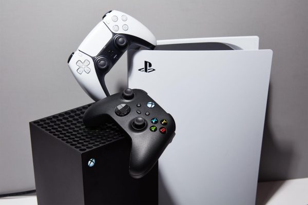 An xbox series x and a PS5 with their controllers defying the laws of gravity to sit weirdly on top of each console.
